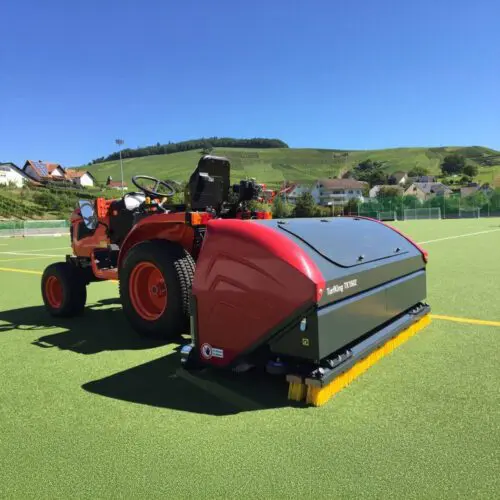 SMG TurfKing TK1502 turf cleaner tractor attachment on soccer field SMG TurfKing TK1502 turf cleaner attached to a tractor on an artificial soccer field with rolling green hills in the background.