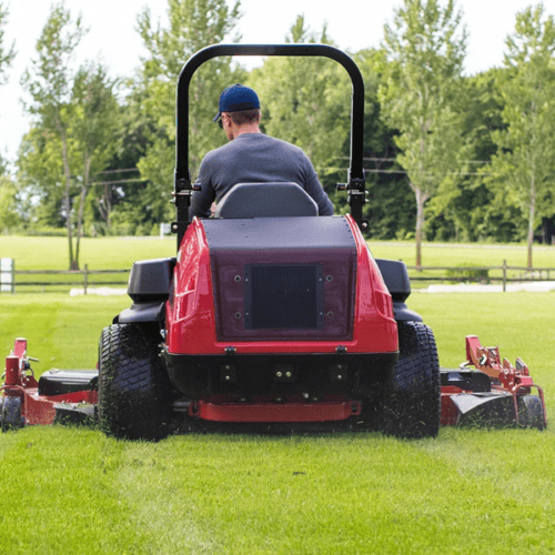 A person operates a red zero turn mower on a grassy field surrounded by trees