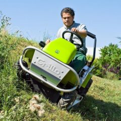 A person operates a green and gray ride-on mower on a sloped grassy area