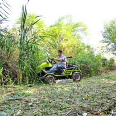 A person operates a green ride-on mower in dense vegetation, showcasing its powerful cutting ability