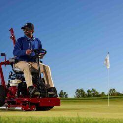 A person operates a red ride-on greens roller on a golf course, with a flag in the background