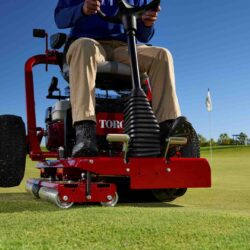 A person operates a red ride-on greens roller on a golf course, with a clear blue sky above