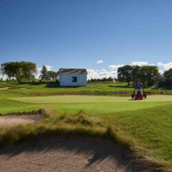 A golfer operates the Toro GreensPro 1260 on a lush green golf course under a clear blue sky