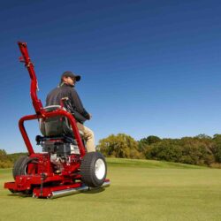 A person operates a red ride-on greens roller on a golf course under a clear blue sky