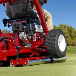 A close-up of a red ride-on greens roller on a golf course, showcasing its drive roller and operator