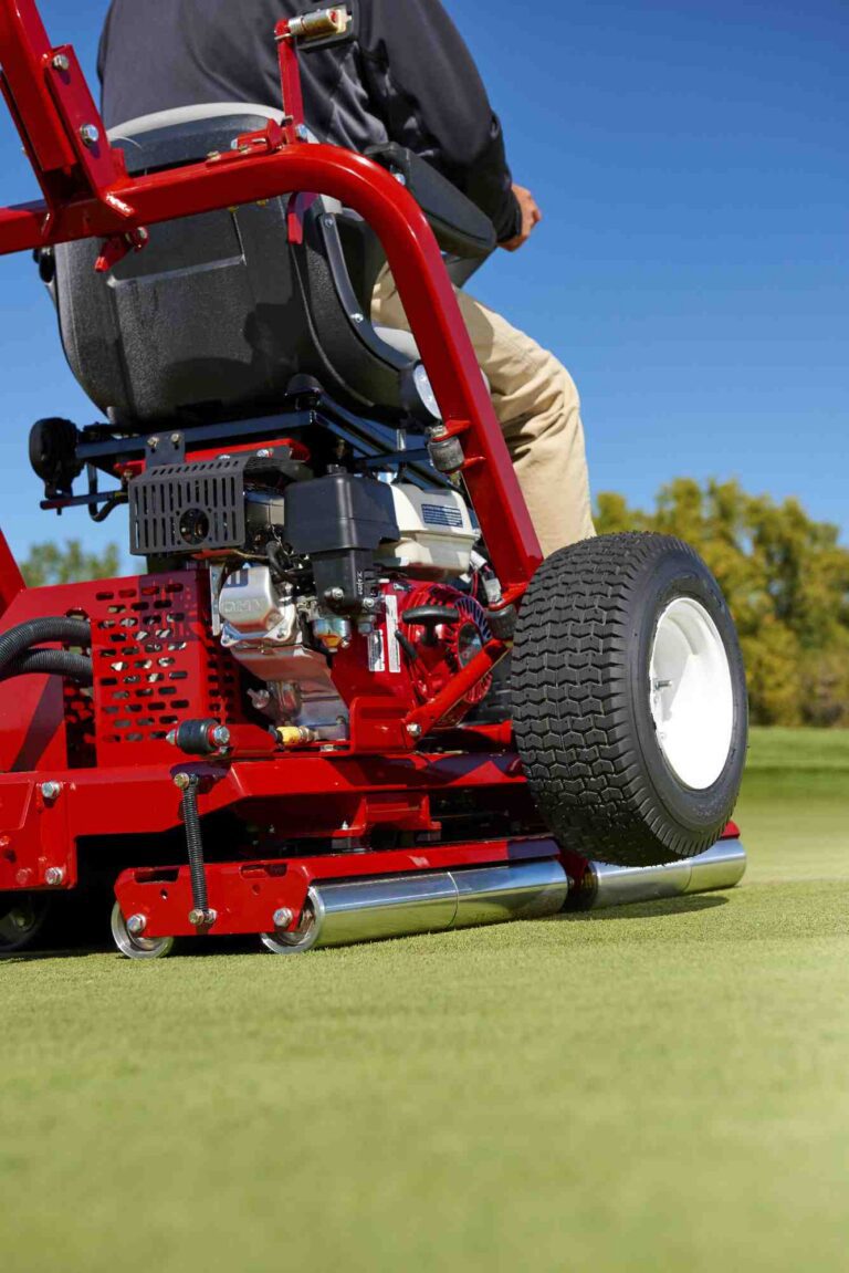 A close-up of a red ride-on greens roller on a golf course, showcasing its drive roller and operator
