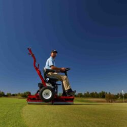 A person operates a red ride-on greens roller on a golf course under a clear blue sky