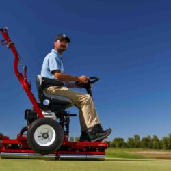A man operates a red ride-on greens roller on a golf course under a clear blue sky