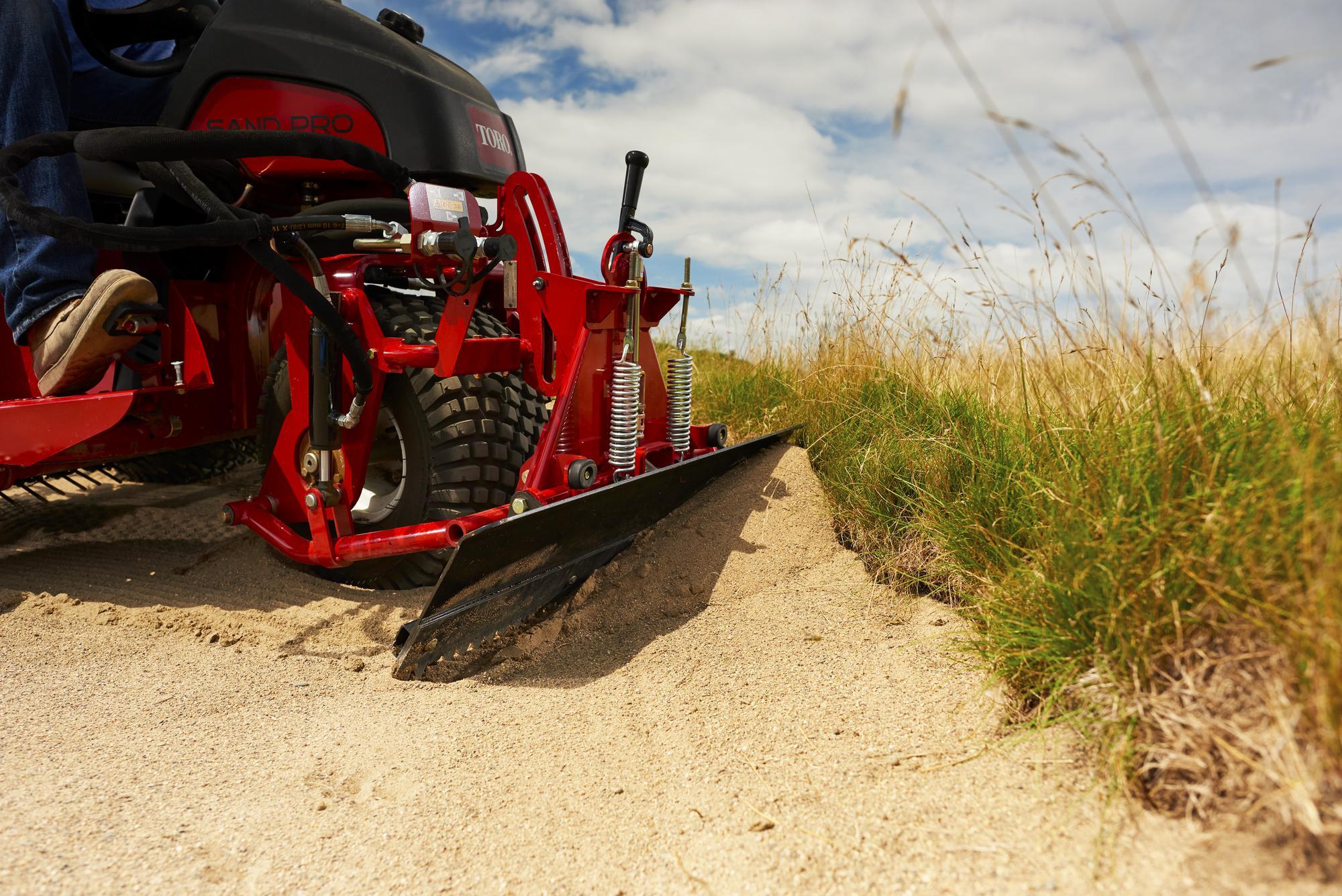 A red mechanical rake grooming sand, showcasing its zero-turn capability in a grassy area