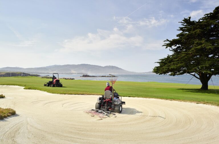 Two sand rakes working on a golf course bunker with a scenic coastal background and green grass