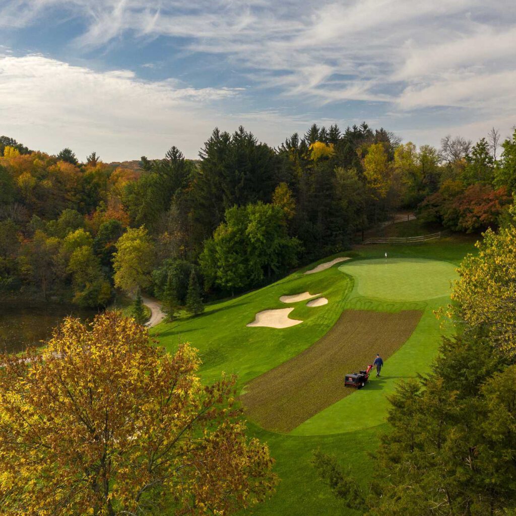 Aerial view of a turf aerator working on a green, surrounded by trees and autumn foliage