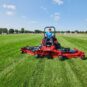 A person operates a red Toro zero turn mower on a grassy field under a clear blue sky