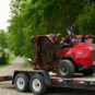 A red Toro zero turn mower on a trailer, with a person driving, surrounded by greenery