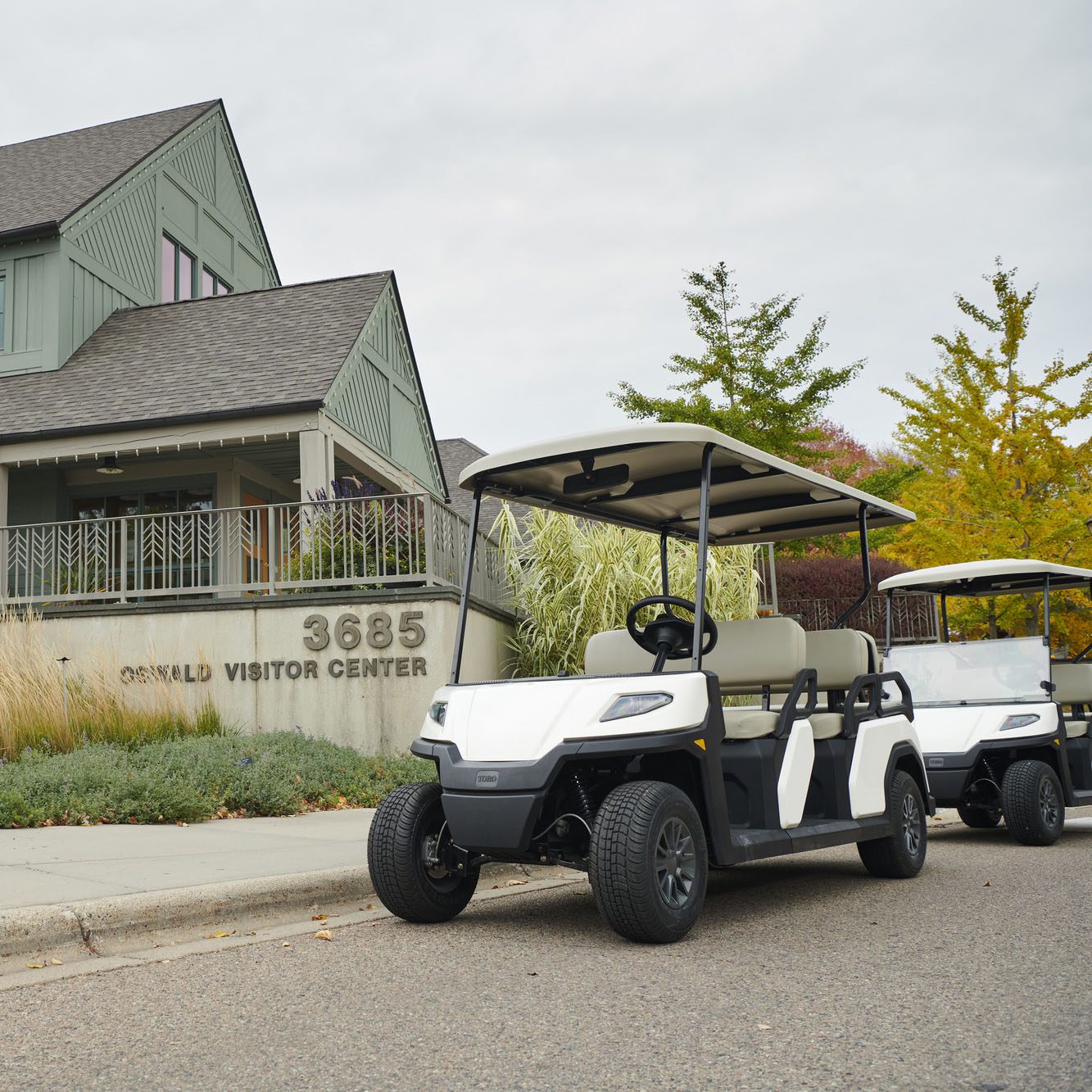 Toro Vista 4-seater golf cart parked at Oswald Visitor Center with autumn trees Toro Vista 4-seater golf cart with beige seats and a white roof, parked outside the Oswald Visitor Center, with another golf cart in the background