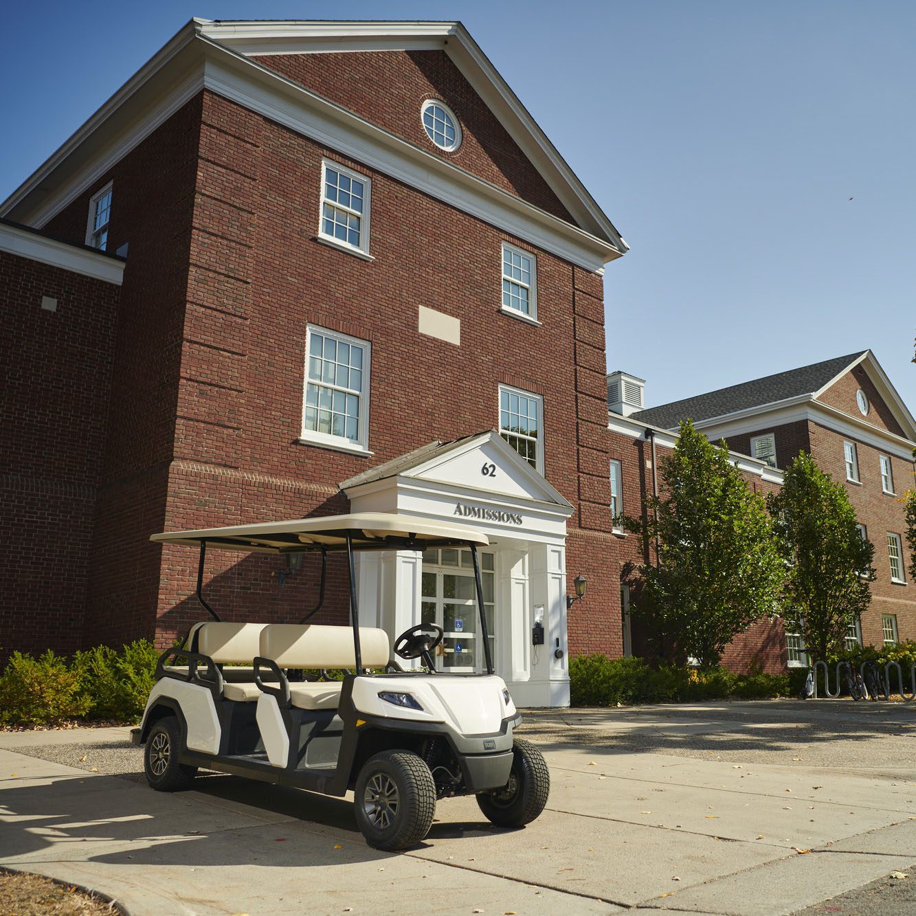 Toro Vista 6-seater golf cart parked outside brick admissions building Toro Vista 6-seater golf cart with beige seats, parked outside a university admissions building, surrounded by brick architecture and greenery