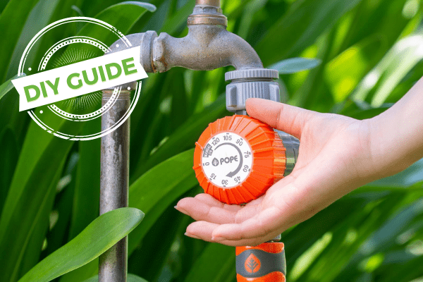 A hand adjusts a garden hose timer near a faucet surrounded by green plants