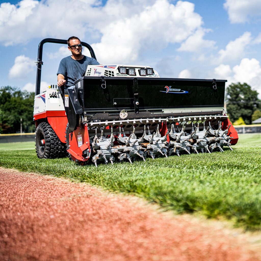 A tractor attachment aerates turf with multiple tines, operated by a person on a grassy field