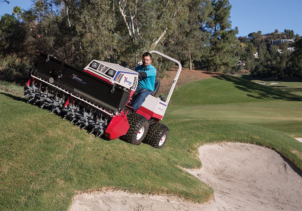 A person operates a red and white tractor attachment on a golf course, stirring soil without damaging turf