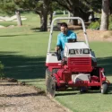 A man operates a red tractor attachment, edging a flower bed on a golf course
