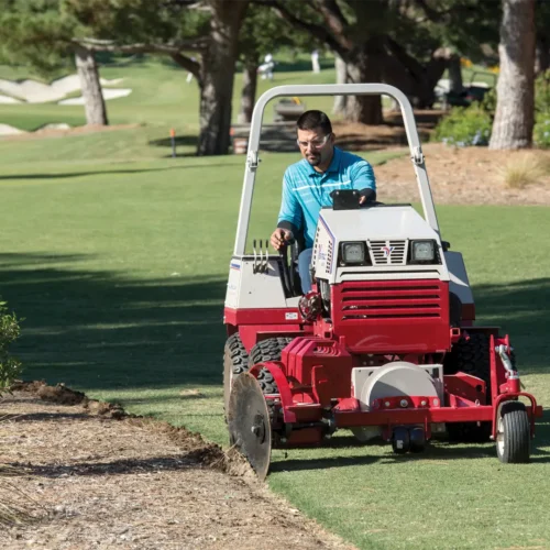 A man operates a red tractor attachment, edging a flower bed on a golf course