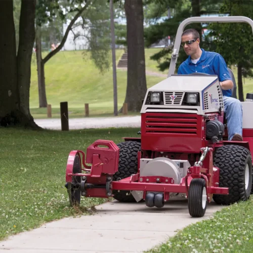 A person operates a red tractor attachment edging a sidewalk in a grassy area