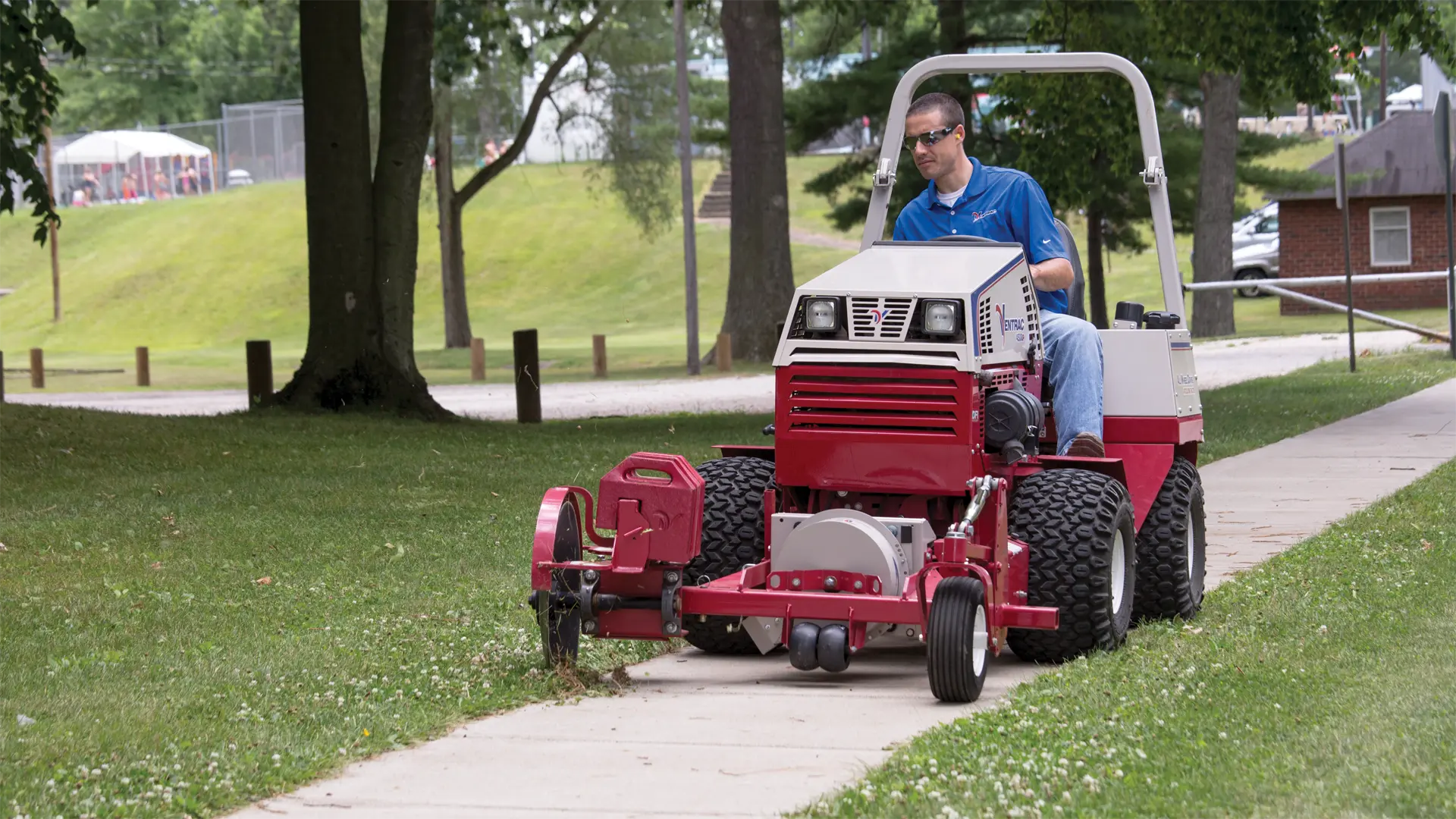 ED200_OrrPark_8992-L1920 A person operates a red tractor attachment edging a sidewalk in a grassy area