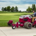 A red tractor attachment edges a sidewalk near a golf course, with a seated operator