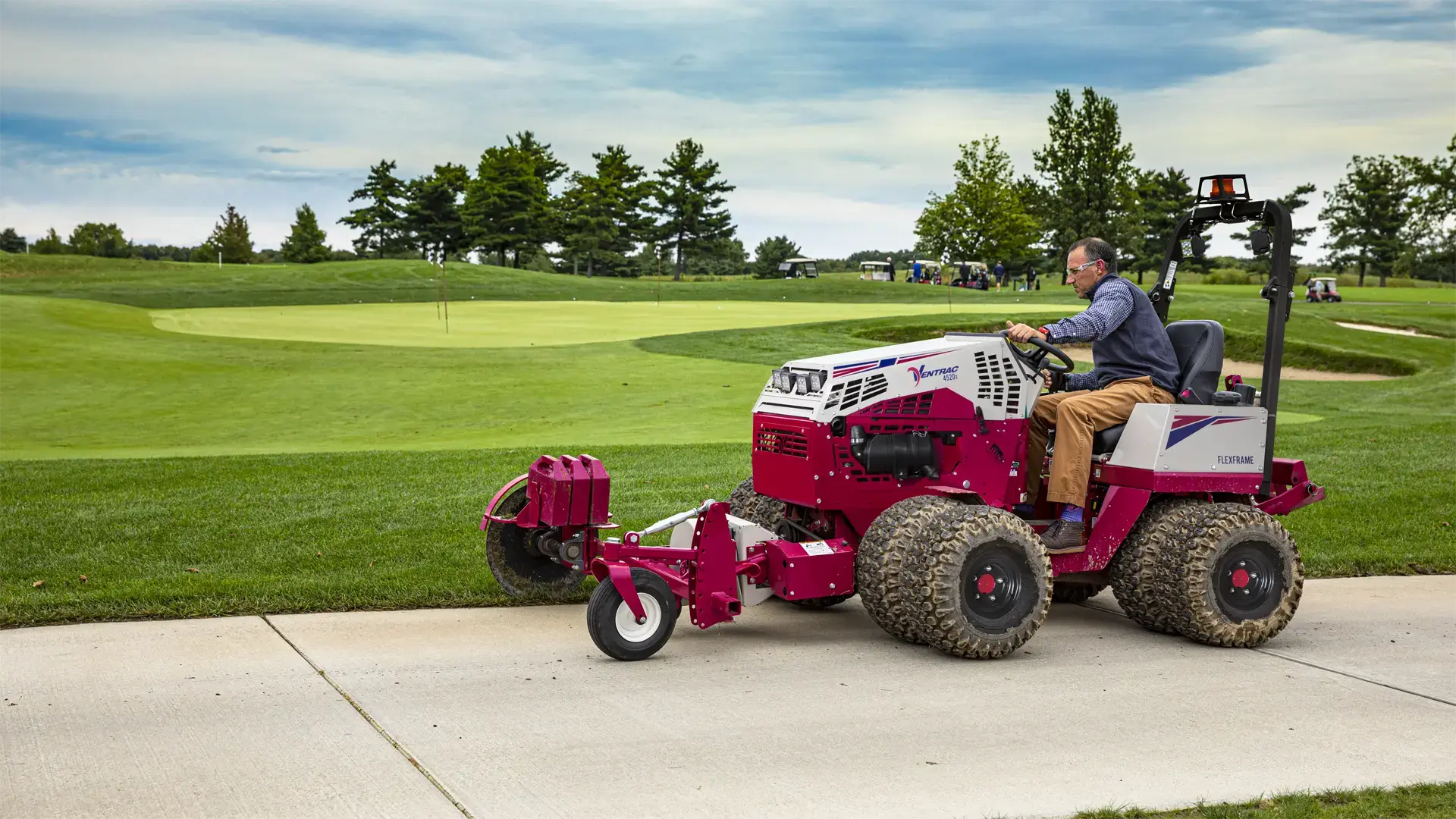 ED200_Westfield-Demo_092718_0911_4520-L1920 A red tractor attachment edges a sidewalk near a golf course, with a seated operator