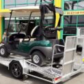A green golf cart on a low-deck trailer with a light ramp and mag wheels outside a building
