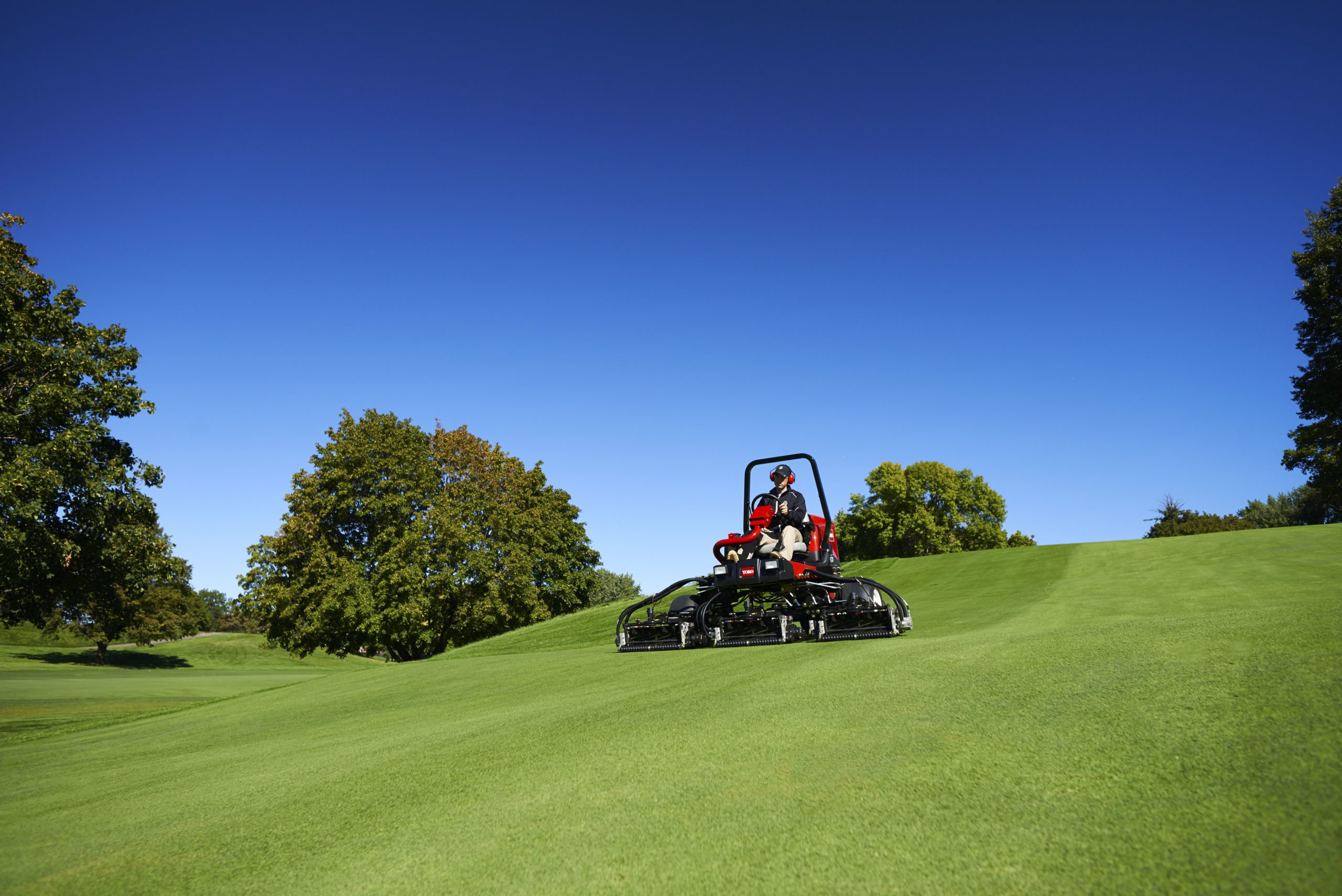 A mower operates on a lush green golf course under a clear blue sky, showcasing premium turf management
