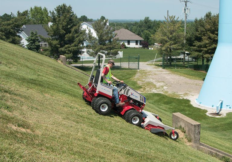 A person operates a red and white tractor on a grassy slope near residential buildings and a water tower