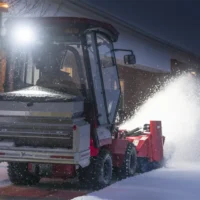 A tractor attachment clears snow from a walkway, illuminated by a bright front light at night