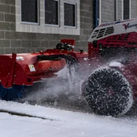 A red tractor attachment sweeping snow from a walkway, showcasing its brush and operational design