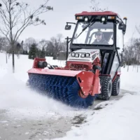 A red tractor with a blue broom attachment clears snow from a walkway in a snowy landscape