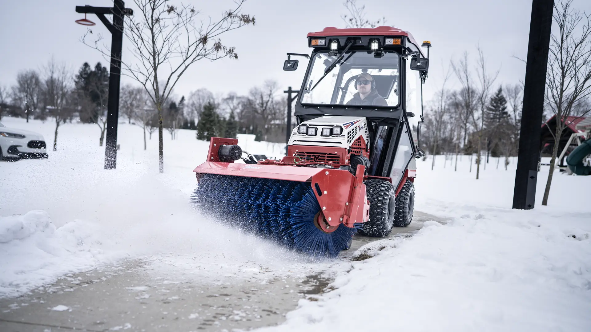 KJ520_P_Graham_Dunn_01296-L1920 A red tractor with a blue broom attachment clears snow from a walkway in a snowy landscape