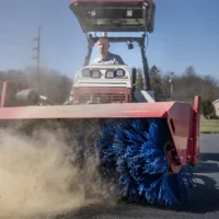 A tractor with a blue broom attachment sweeps dirt on a sunny day, showcasing its cleaning capability