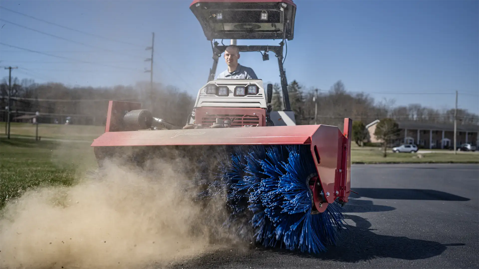 KJ_Broom_Orrville_School_4146-L1920 (3) A tractor with a blue broom attachment sweeps dirt on a sunny day, showcasing its cleaning capability