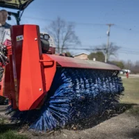 A red broom attachment on a tractor sweeps dirt and debris from a grassy area