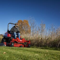 A person operates a red zero-turn mower on a grassy area with trees in the background