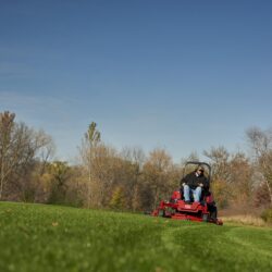 A person operates a red zero turn mower on a grassy field with trees in the background