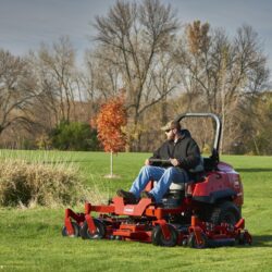 A man operates a red zero turn mower on a grassy field with trees in the background