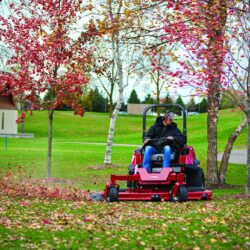 A person operates a red zero turn mower among colorful autumn trees and fallen leaves