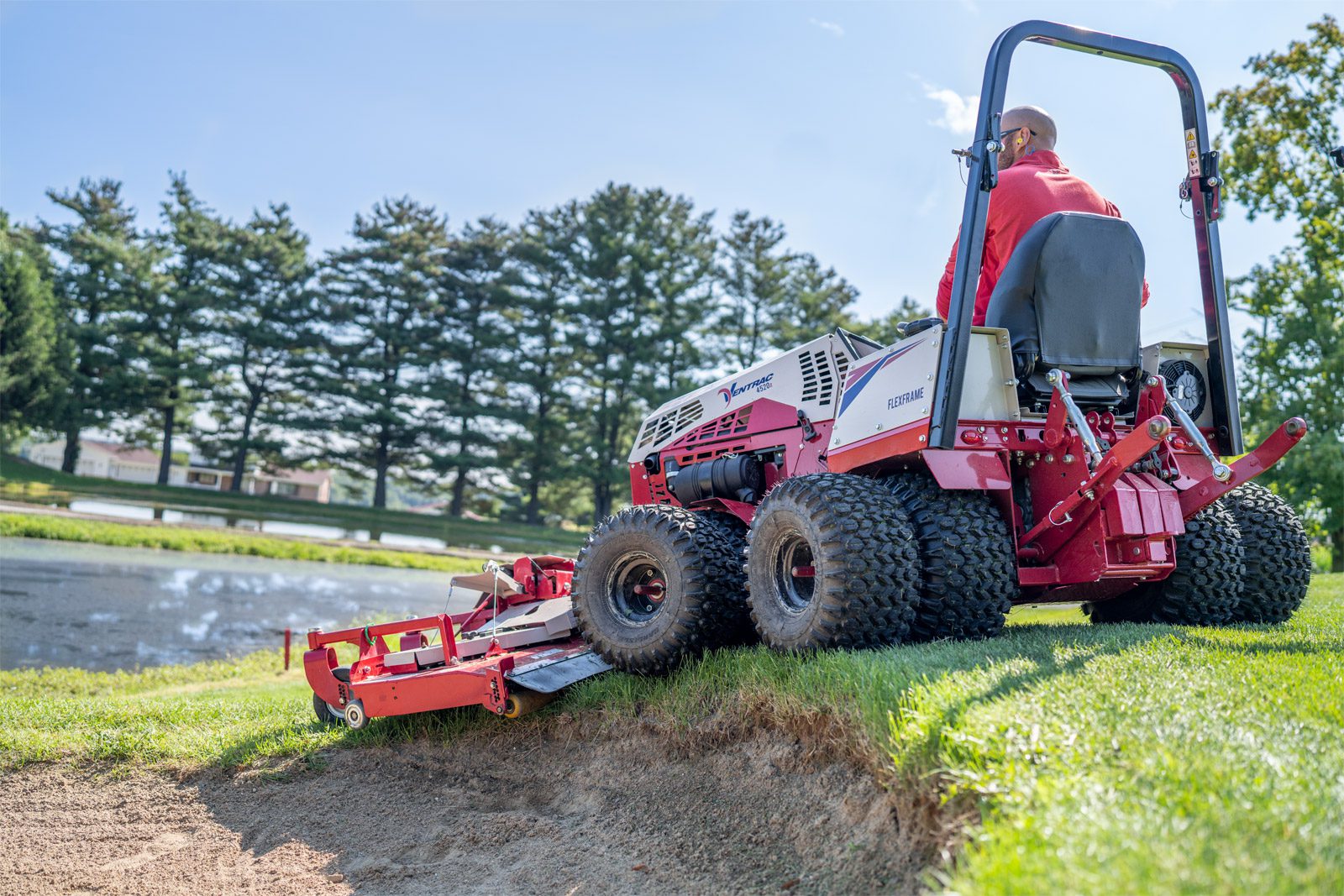 A red Ventrac machine trims grass near a pond on a golf course, showcasing its low profile design