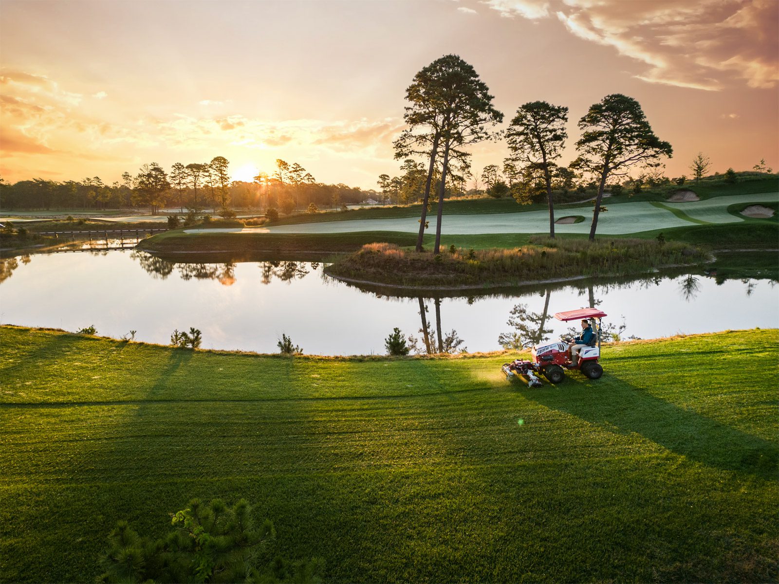A Ventrac machine mows grass near a lake on a golf course at sunset, surrounded by trees