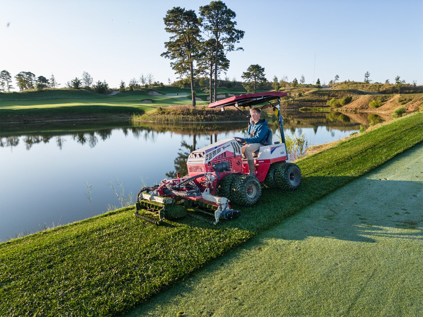 A red Ventrac machine mows grass near a golf course pond under clear blue skies