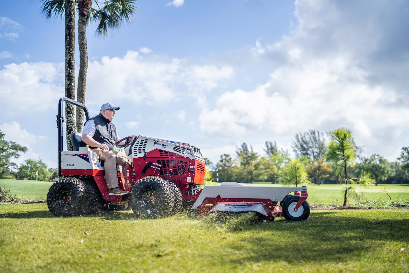 A Ventrac machine mows grass on a golf course, with a user seated and palm trees in the background