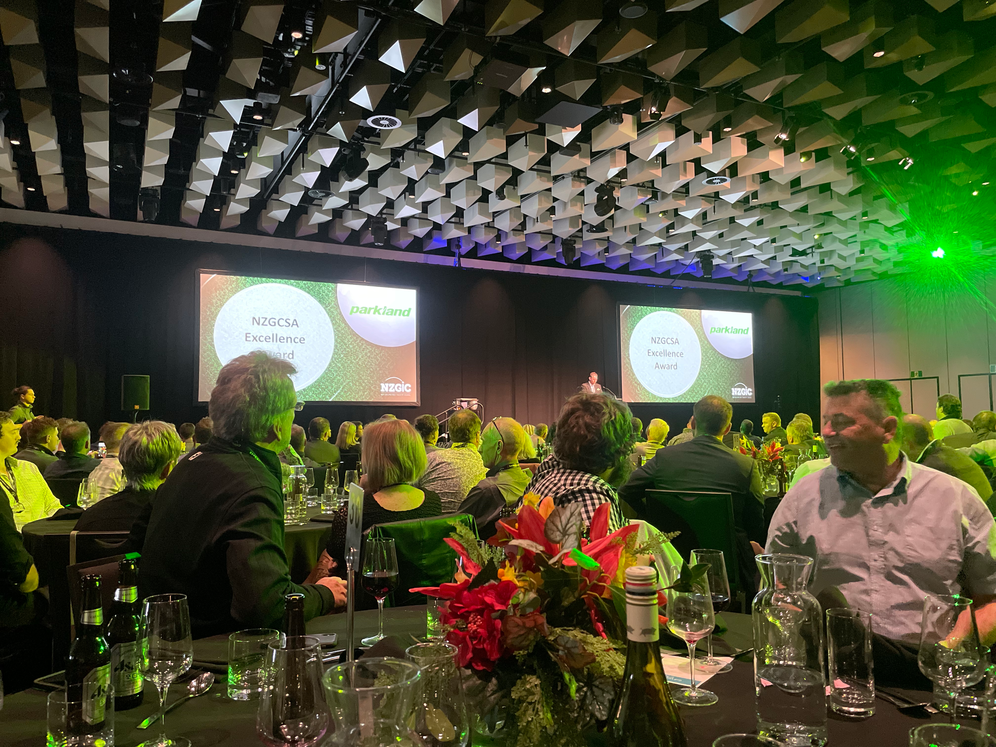 A crowd gathers in a banquet hall for the NZGCSA Excellence Award ceremony, with green lighting and presentations visible