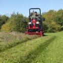 Red ride-on mower in action, cutting grass with a driver, surrounded by greenery and trees