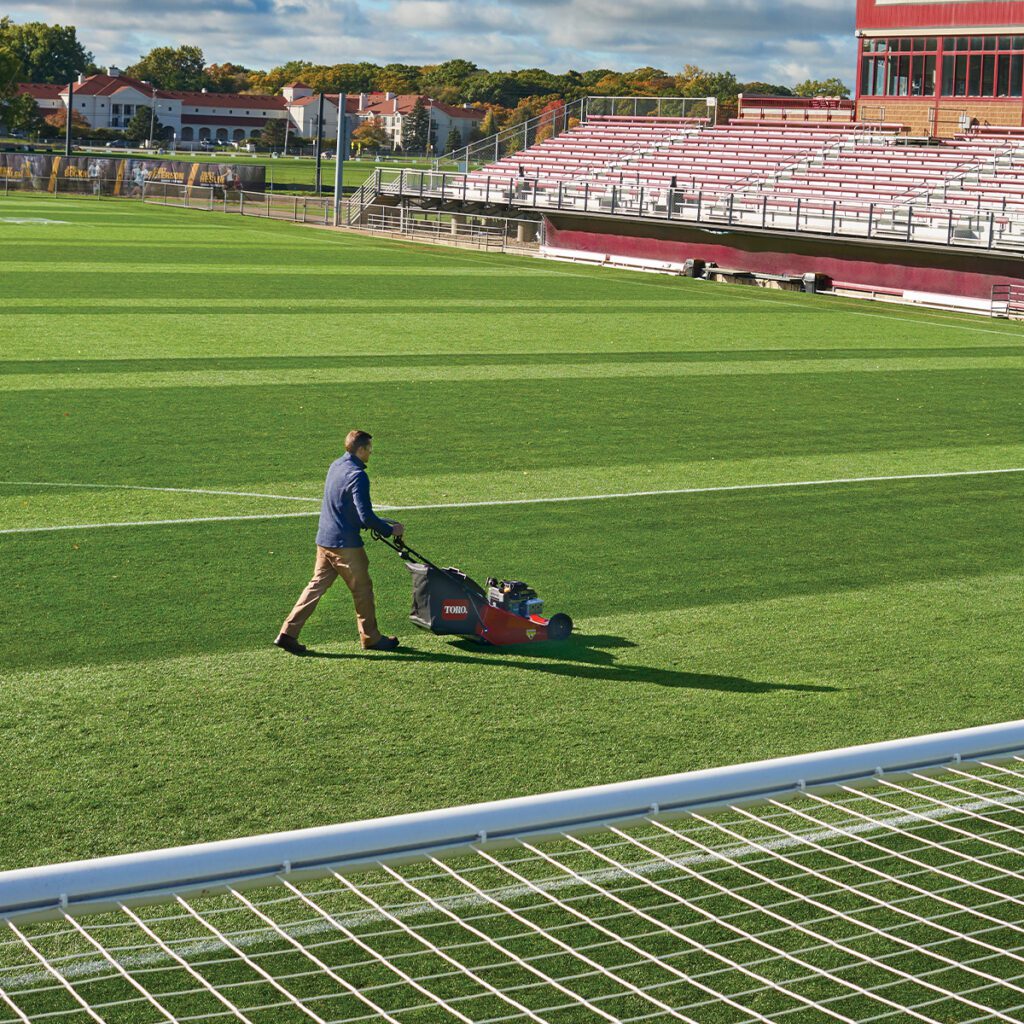 A person operates a Toro mower on a well-maintained sports field with vibrant green grass
