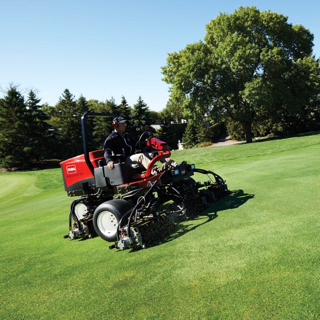 Red ride-on mower in action on a lush green field, showcasing its cutting reels and operator